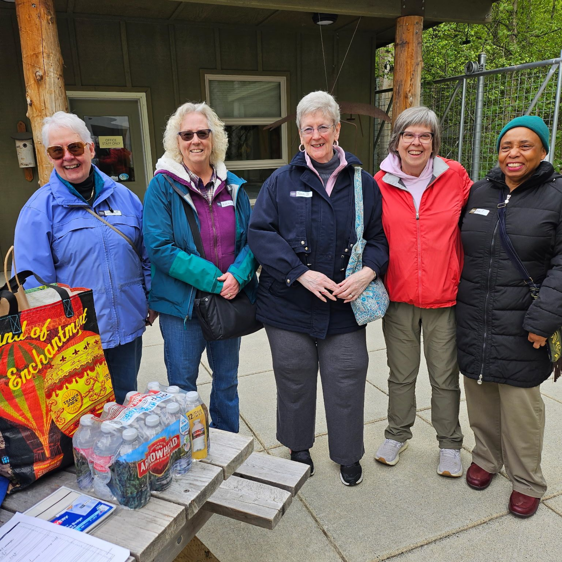Five people standing outdoors, smiling. They stand near a table with bottled drinks.