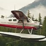 Seaplane in flight; white, red, and silver fuselage; mountain backdrop.