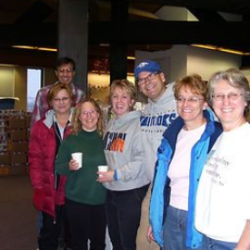 Group of people smiling, posing together indoors, some holding cups, with a storage area in the background.