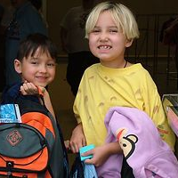 Two children smiling with backpacks. One wears yellow, the other is dark-haired with a blue shirt.