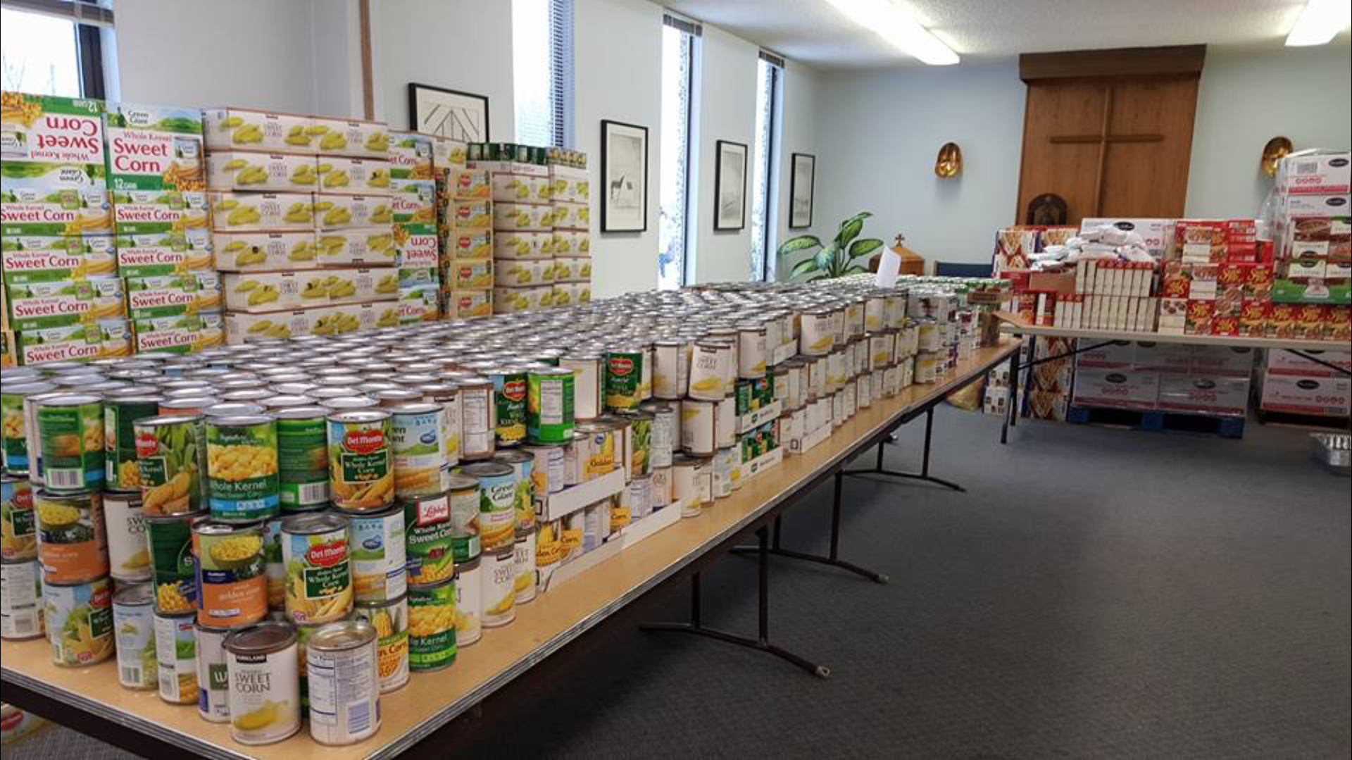Food bank: Canned goods stacked on tables and in boxes; produce visible; neutral-toned room; natural light.
