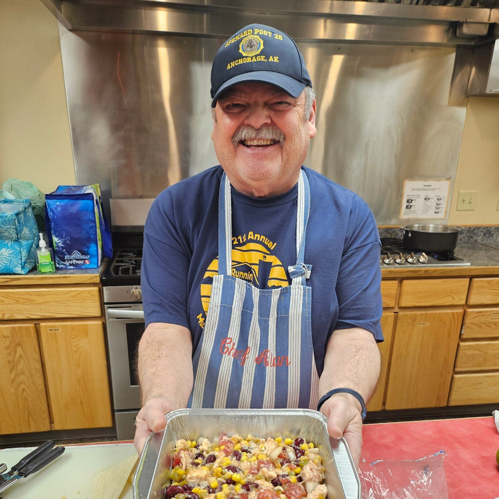 Man in apron holding a pan of food in a kitchen, smiling.