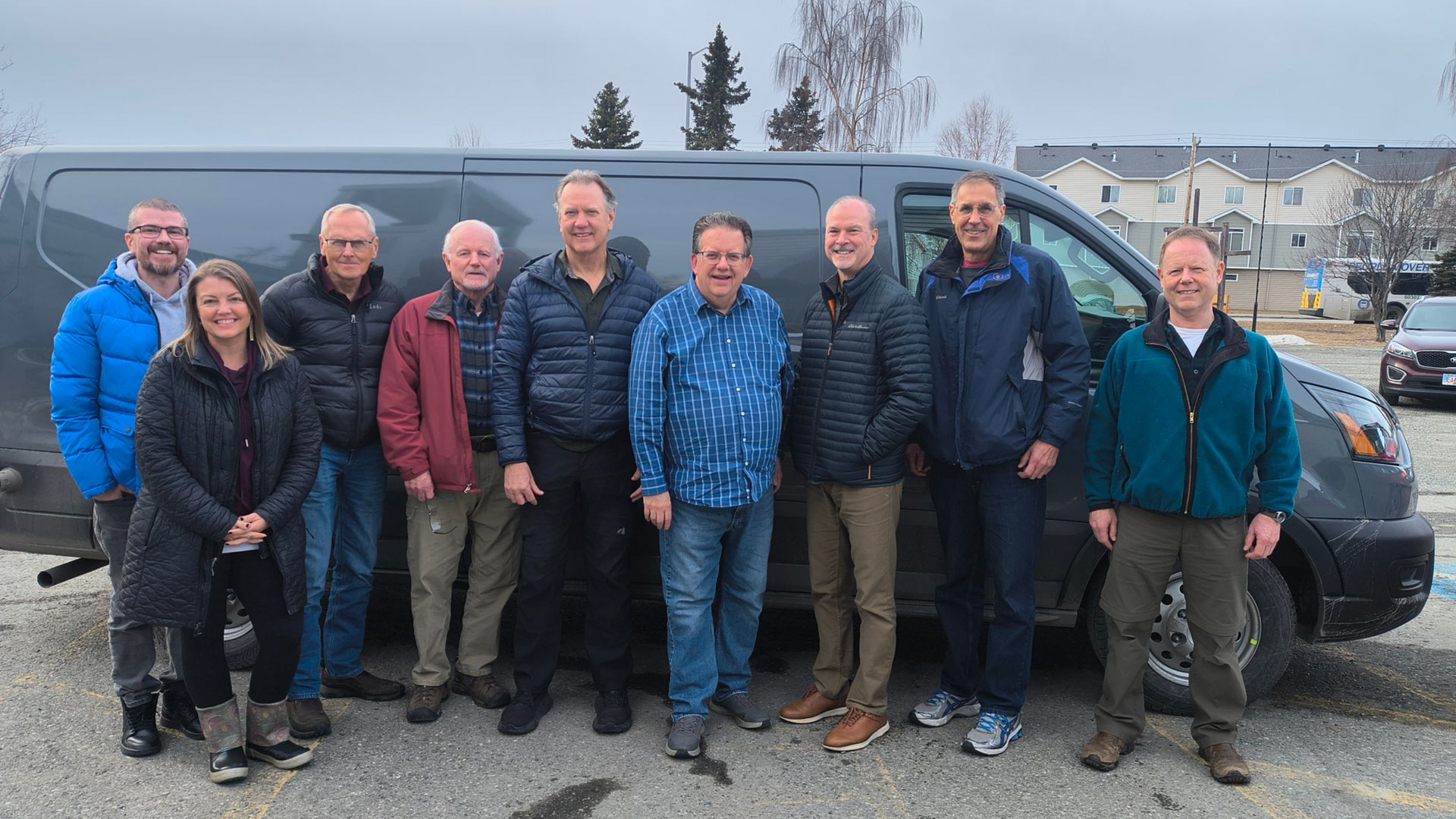 Group of people standing in front of a gray van on a cloudy day, smiling.