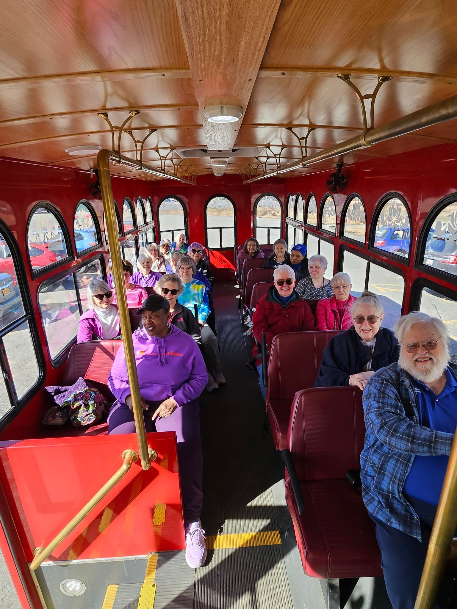 People on a red trolley, looking out windows. Interior view, sunlight, seated passengers.