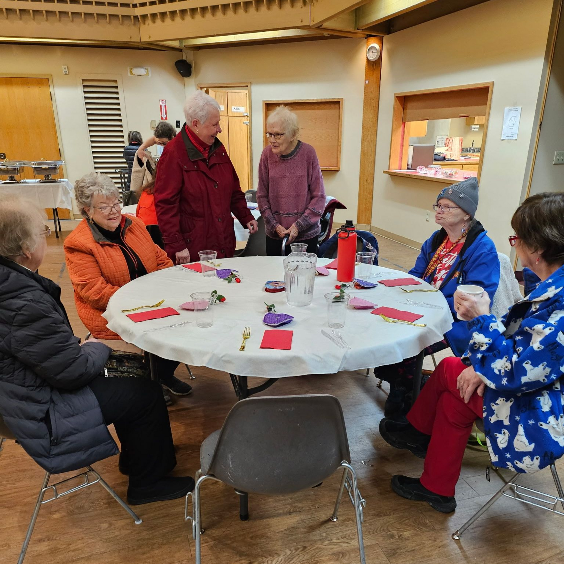 Group of older adults gather around a round table, chatting. Several are wearing coats. Indoor setting with a buffet.