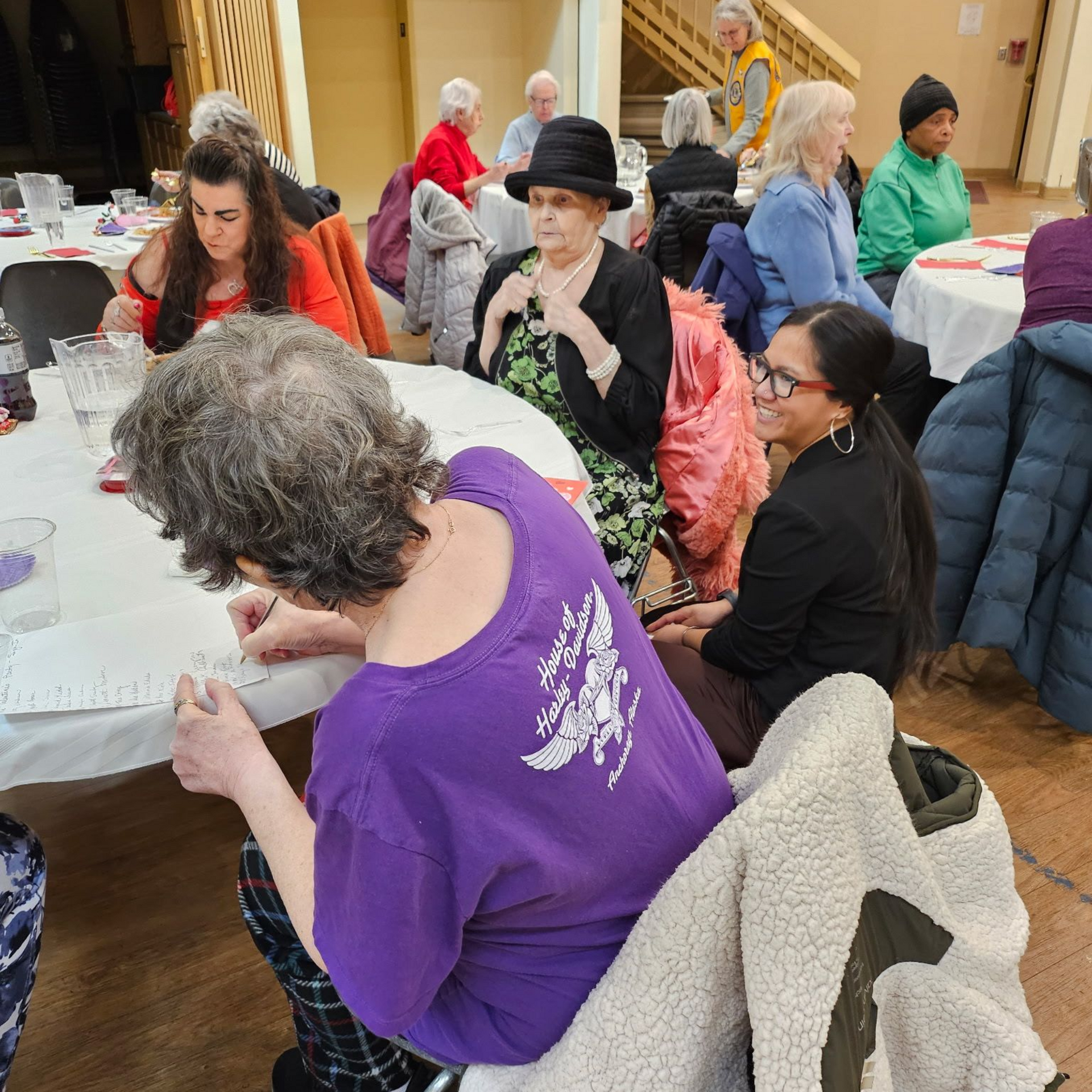 People seated at tables in a community hall, some writing. Several wear coats.