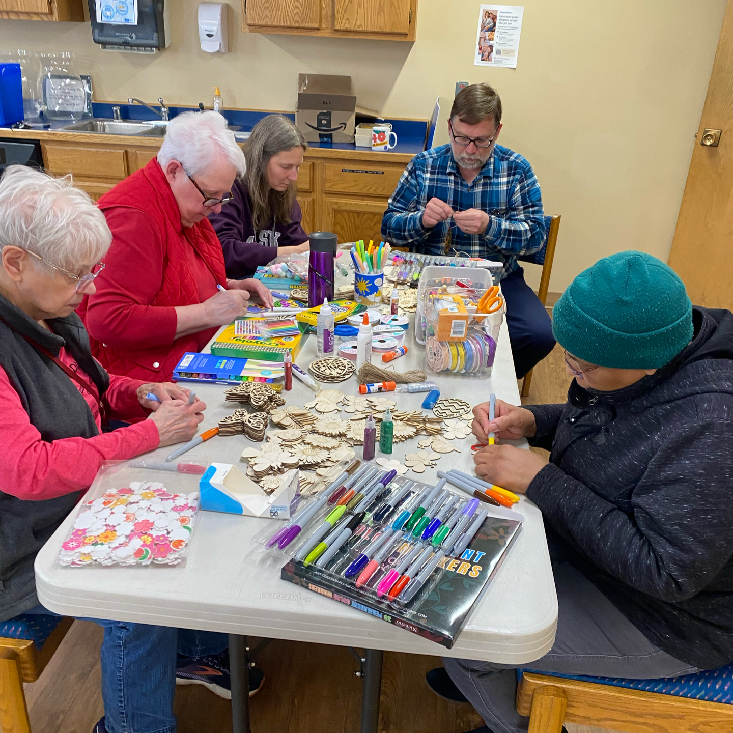 People at a table, crafting with supplies. Colorful markers, wooden shapes, and other items are scattered around.