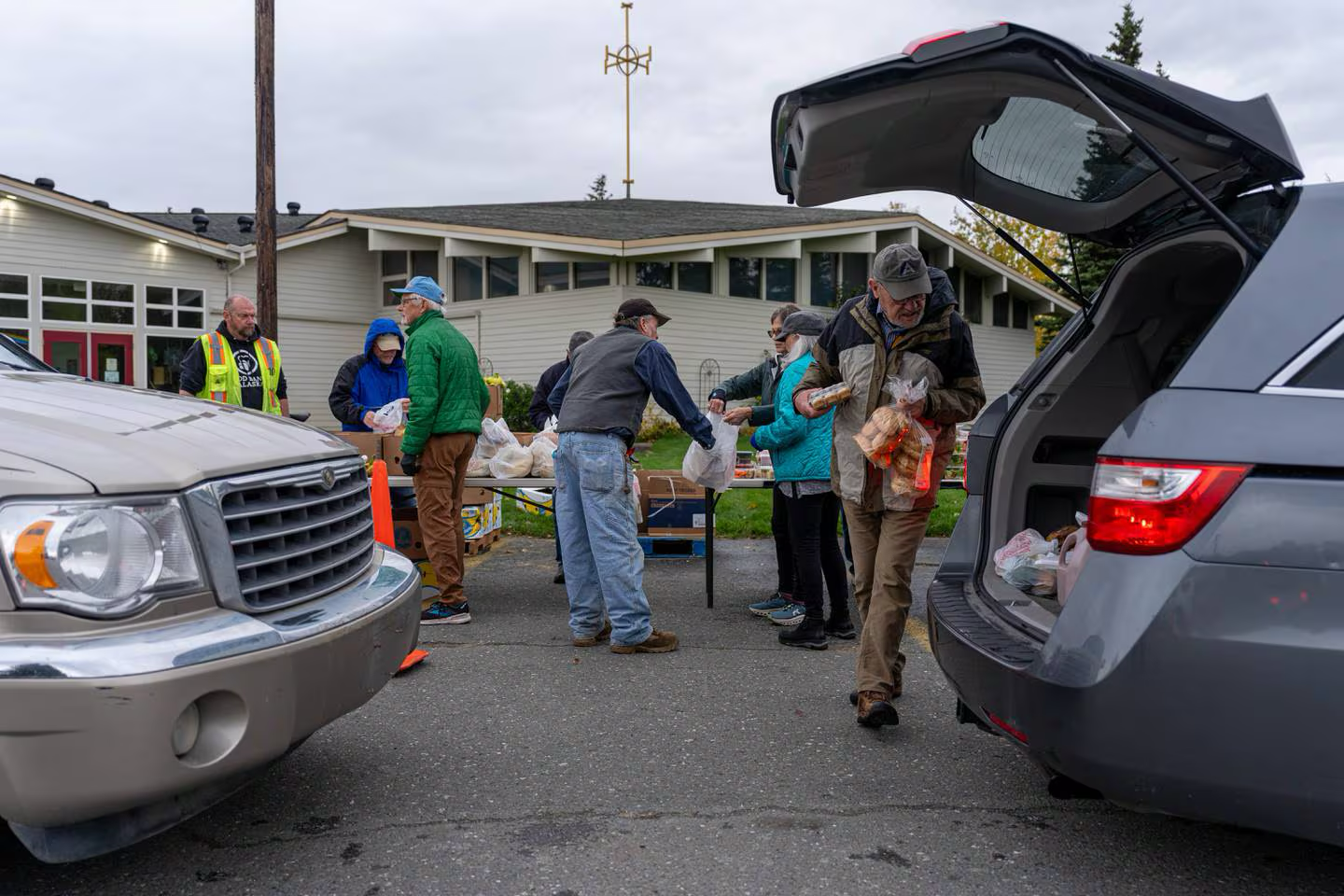 People distributing food from a table and car trunks outside a building.