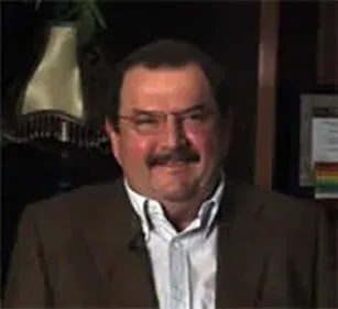 Man in brown jacket, white shirt, and glasses, smiling in an office setting.
