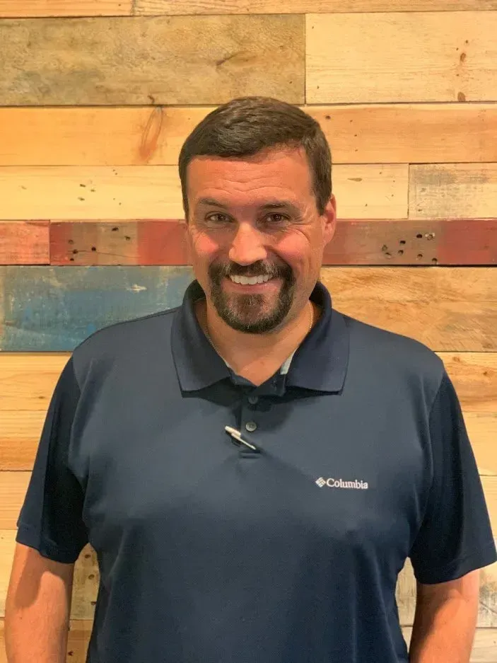 Man with a dark beard wearing a blue polo shirt smiles in front of a wood-paneled wall.