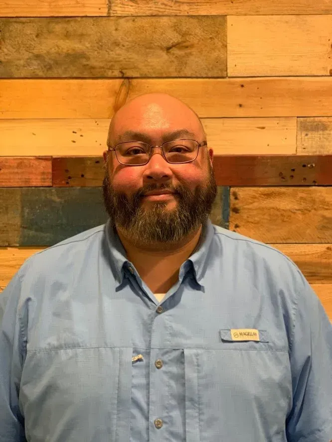 Man with glasses and a beard wearing a light blue button-down shirt, standing in front of a wood panel wall.