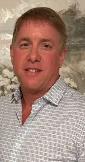 Person in a black blazer and brown button-up shirt smiling. They have curly hair and are indoors against a beige wall.