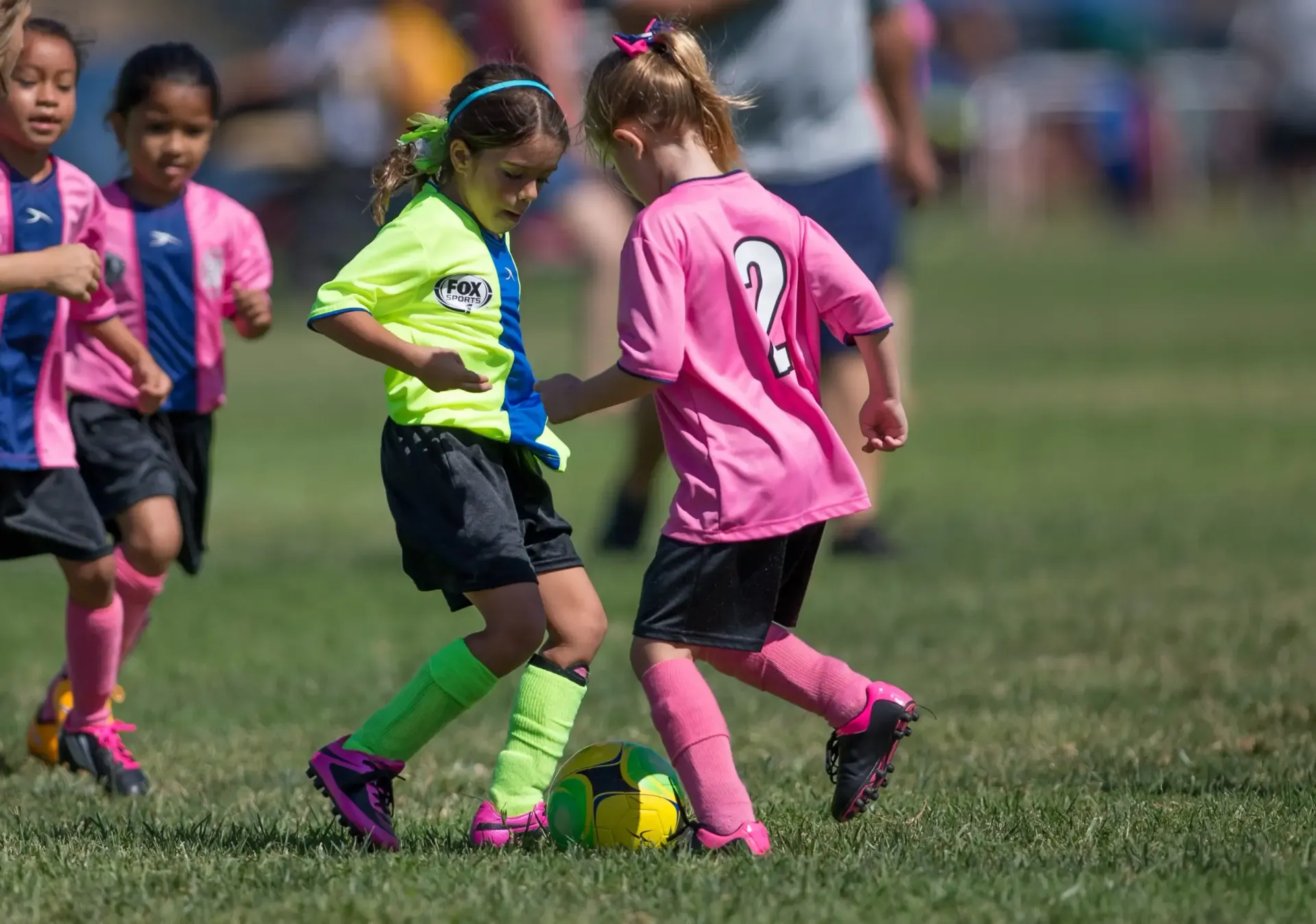 Children playing soccer on a grass field, with one child in a yellow jersey competing for the ball against one in pink.