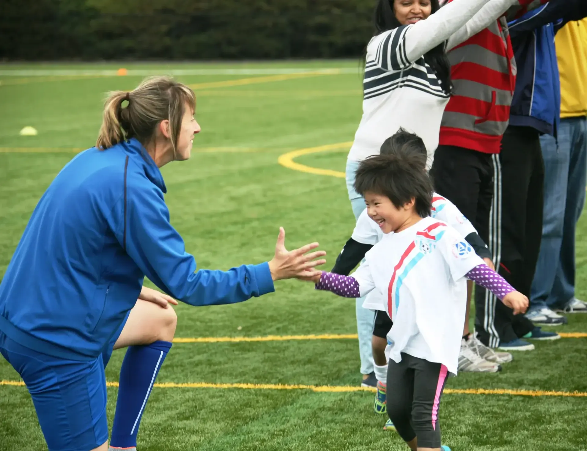A coach in blue athletic gear kneels on a field to high-five a smiling child in a white shirt during an outdoor activity.