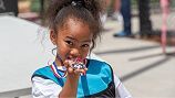 A smiling child wearing a blue sports jersey holds up a small, silver medal near their face, outdoors on a sunny day.