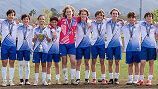 A soccer team in blue and white uniforms stands side-by-side on a grass field, wearing medals and holding a trophy.