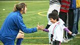 A coach wearing a blue track suit kneels on a field to give a high-five to a child in a white shirt.