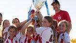 A group of players in red and white striped jerseys hold a championship trophy high while celebrating outdoors.