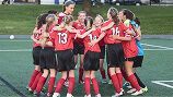 A soccer team in red jerseys and black shorts huddles together on a green turf field.