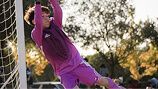 A soccer goalkeeper in a purple uniform jumps toward the goal post to block a shot during a sunny outdoor game.