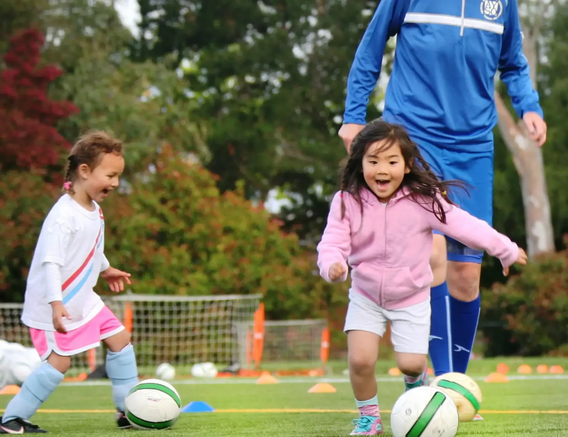Two children in sports clothes joyfully practice soccer on a green field with a coach in the background.