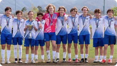 A youth soccer team in blue and white jerseys stands in a line on a field, wearing medals and holding a trophy.