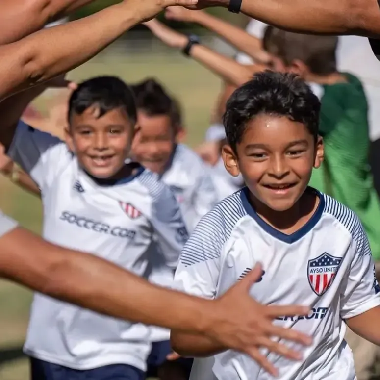 Children in white soccer jerseys run through a high-five tunnel on a grassy field, smiling excitedly.