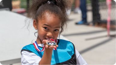 A person in a blue and white sports jersey holds a soccer-themed medal toward the camera with a joyful expression.