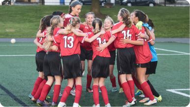 A group of soccer players in red jerseys and black shorts huddle together on a green turf field.