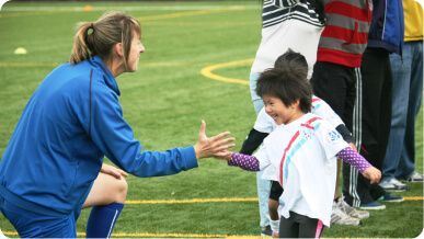 A coach in a blue uniform kneels on a field to give a high-five to a smiling child wearing a white sports shirt.
