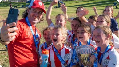 A coach holding a phone to take a selfie with a joyful soccer team celebrating with a trophy on a field.