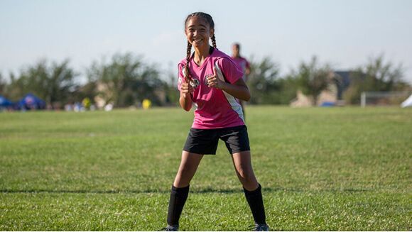 A soccer player in a pink jersey and black shorts stands on a grass field giving a thumbs-up.