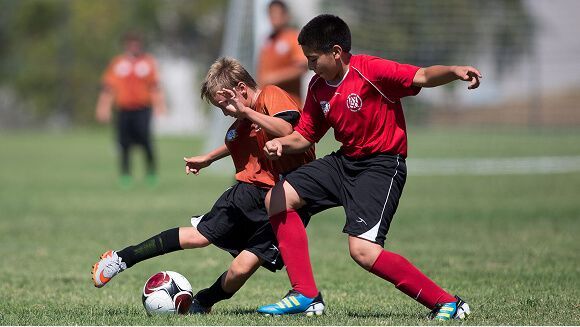 Two young soccer players in red and orange jerseys compete for the ball on a grassy field.