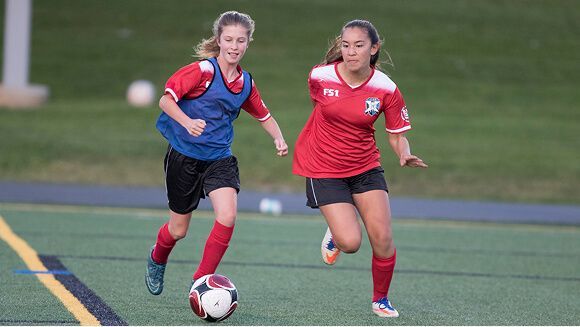 Two soccer players in red jerseys run side-by-side on a turf field, with one player dribbling the ball.