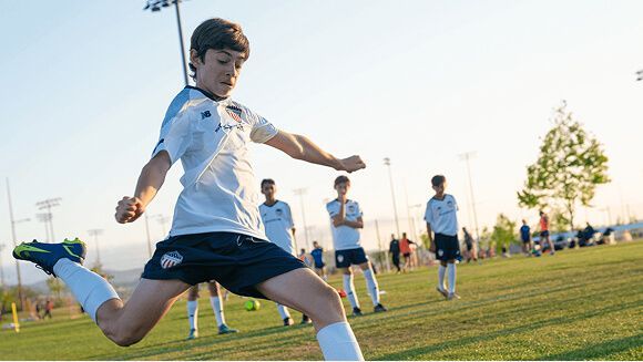A youth soccer player in a white jersey and blue shorts kicks a ball on a sunny grass field with teammates in the distance.