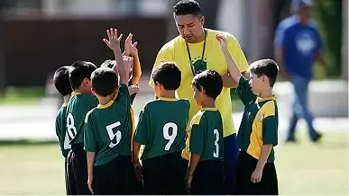 A coach in a yellow shirt gathers a youth soccer team in green uniforms on a field as they put their hands together.