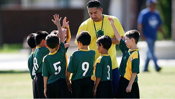 A coach in a yellow shirt stands with a team of children in green and yellow soccer uniforms in a huddle on a field.
