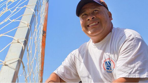 A smiling person in a baseball cap and white t-shirt stands next to a soccer goal post against a clear blue sky.