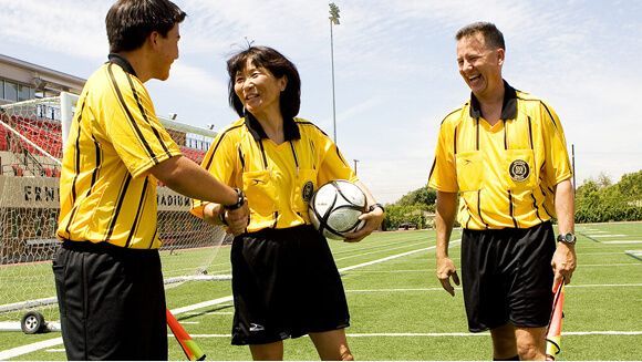 Three soccer referees in matching yellow shirts and black shorts stand on a field, smiling and holding a ball and flags.