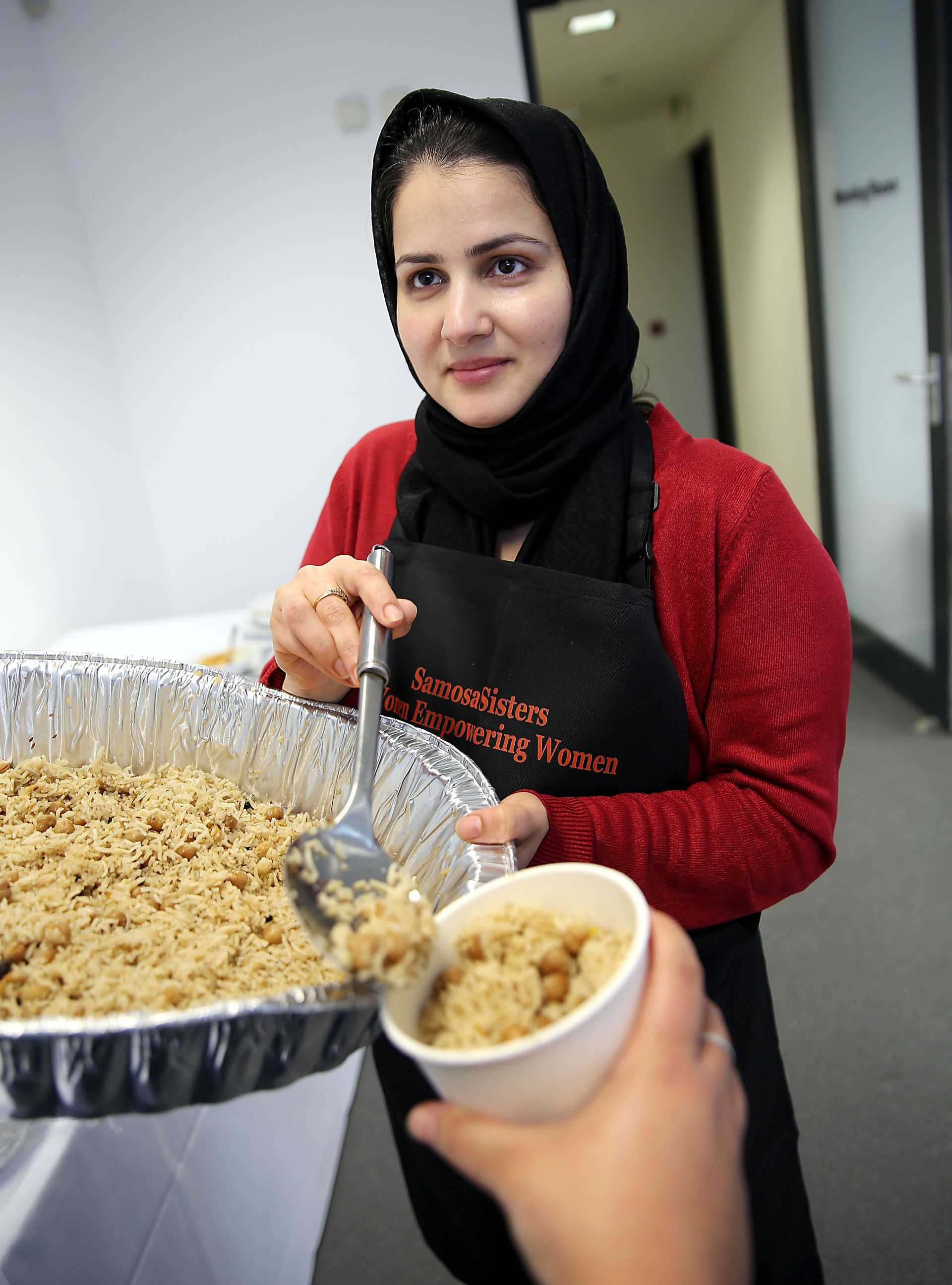 A woman in a red sweater and black apron is pouring food into a bowl
