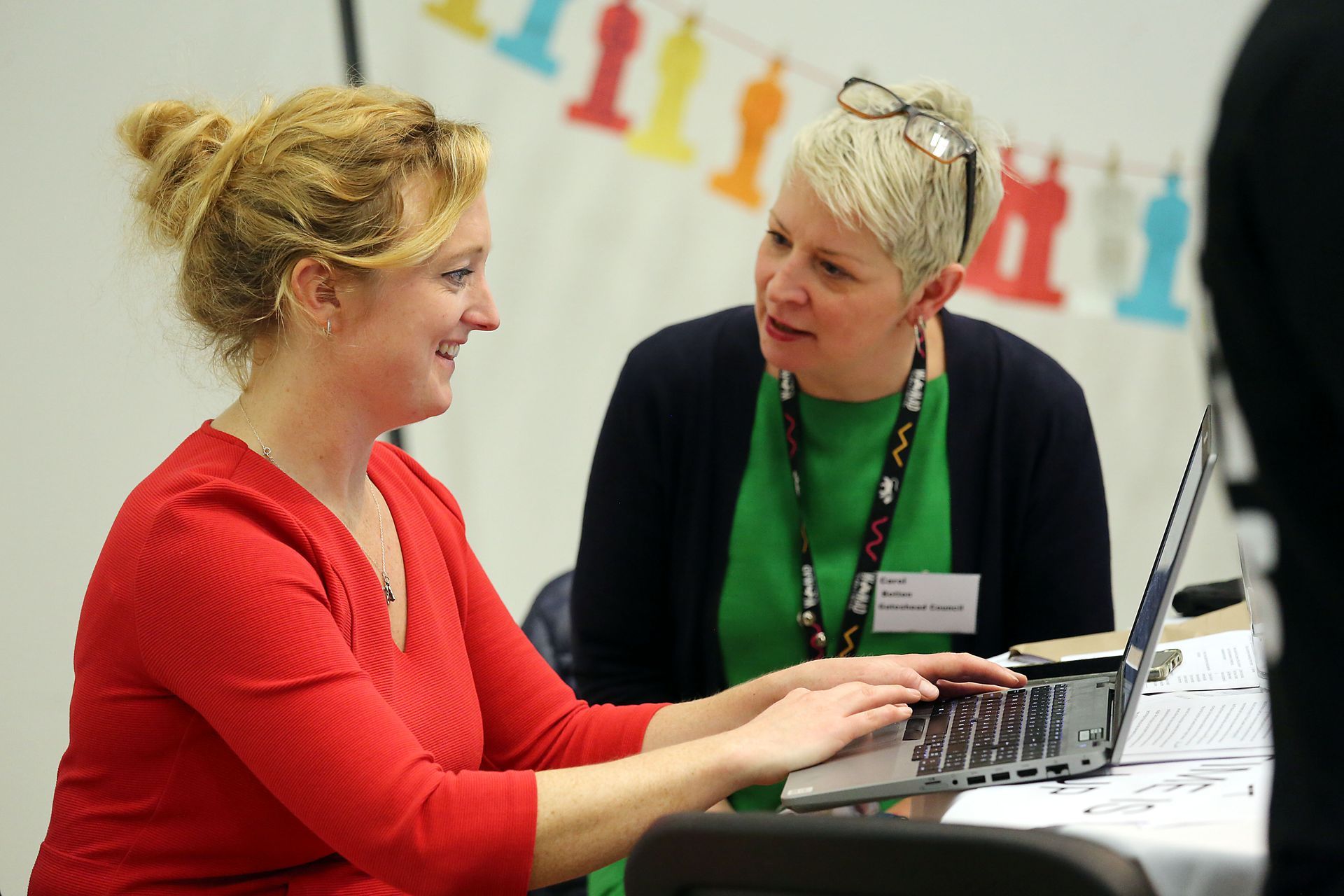 Two women are sitting at a table with a laptop and talking to each other.