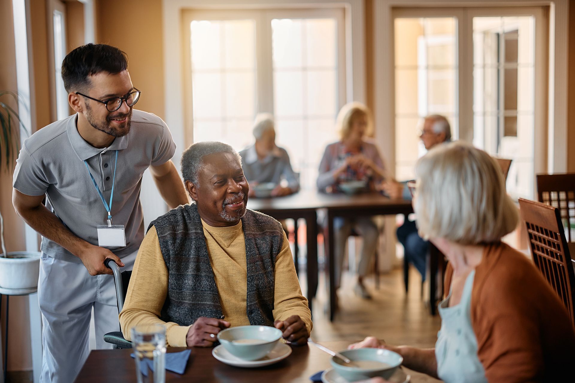 A man is talking to an elderly woman sitting at a table in a nursing home