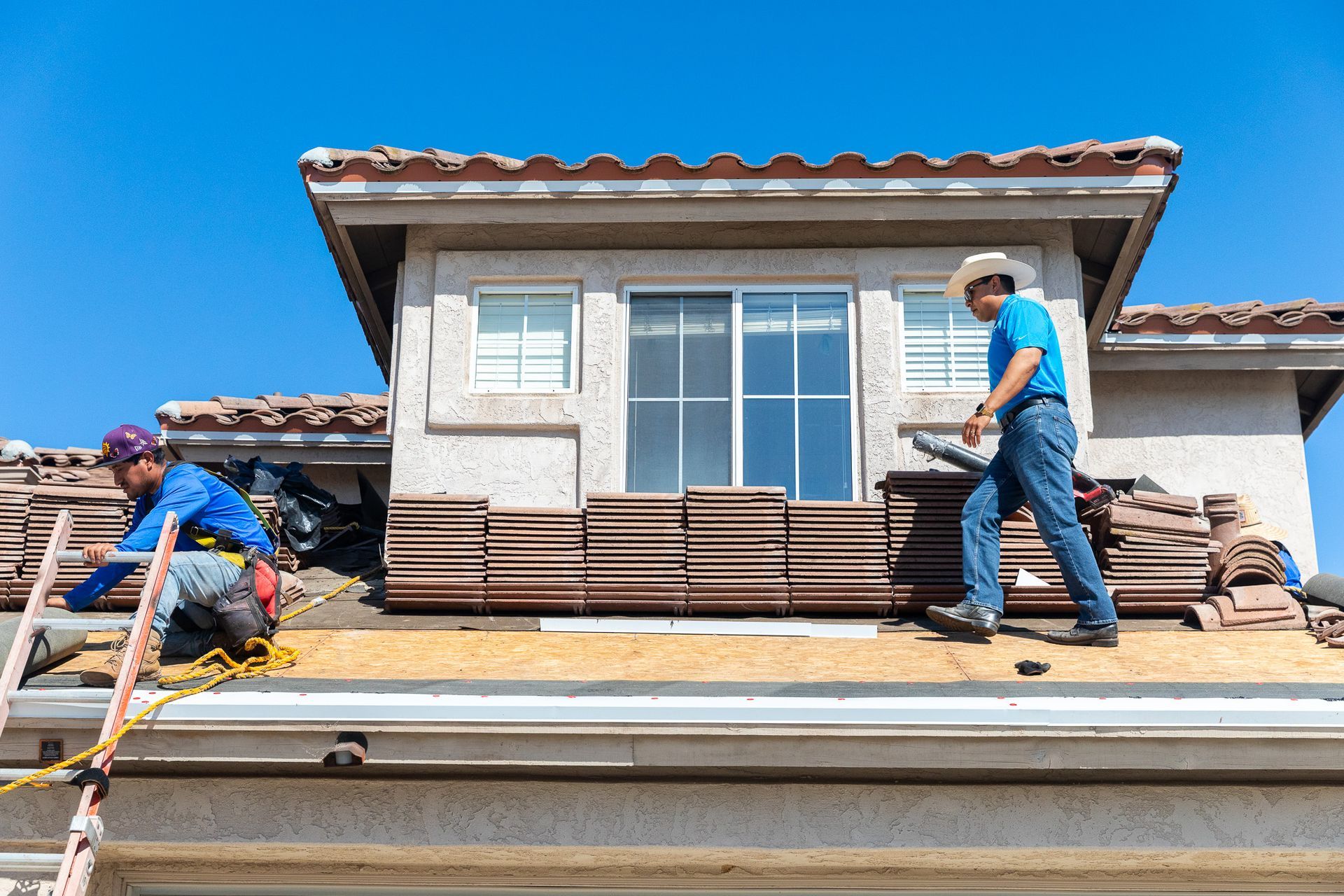 Two roofers in work clothes and hats replace tiles on a residential home roof under a clear blue sky.