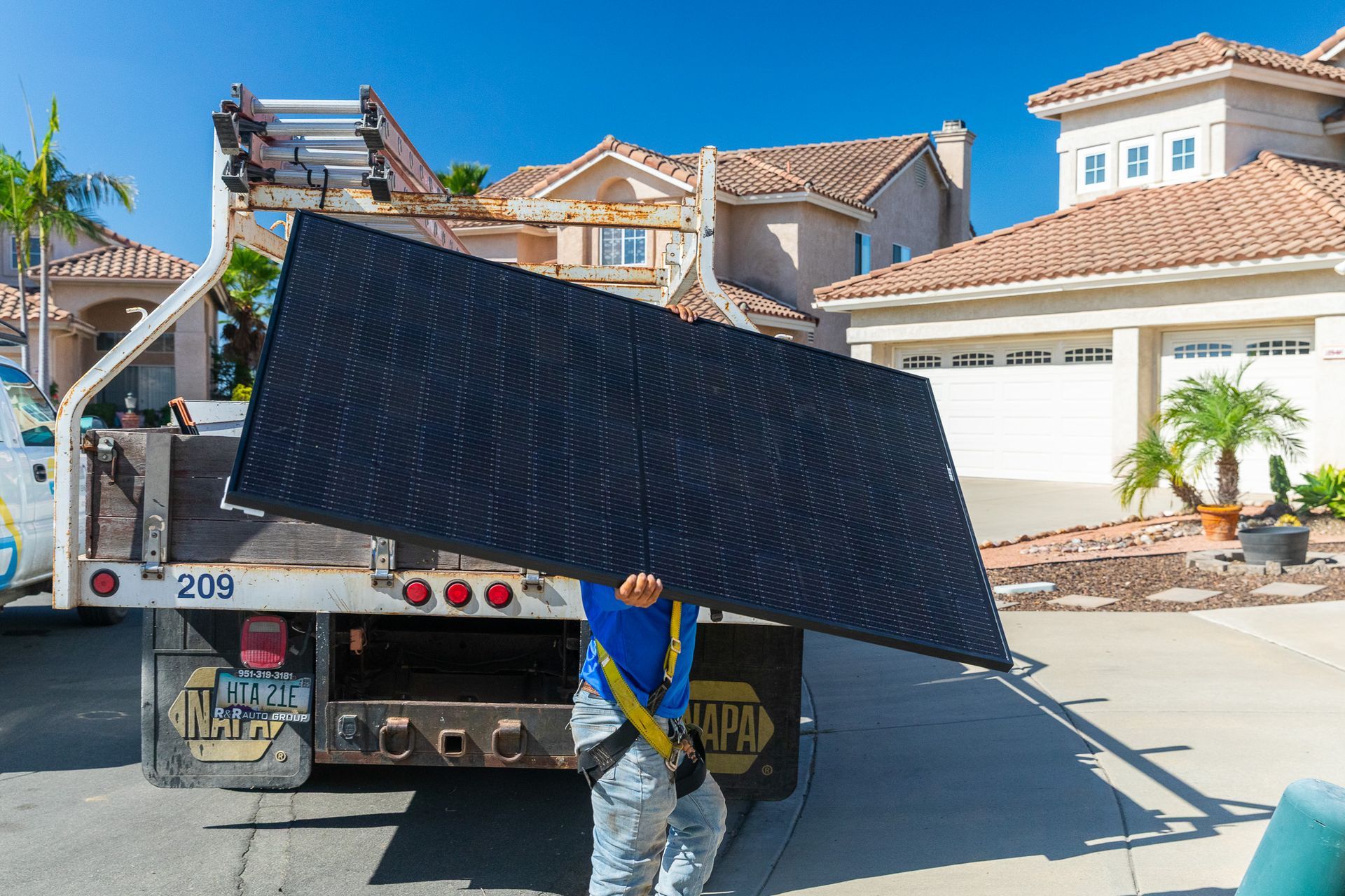 Worker holding a large black solar panel near a truck in front of houses on a sunny day.