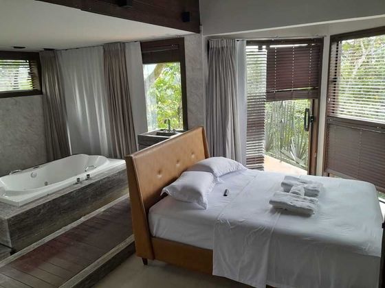 Um quarto com cama de cabeceira de couro e banheira de imersão branca ao lado de janelas do chão ao teto com persianas.