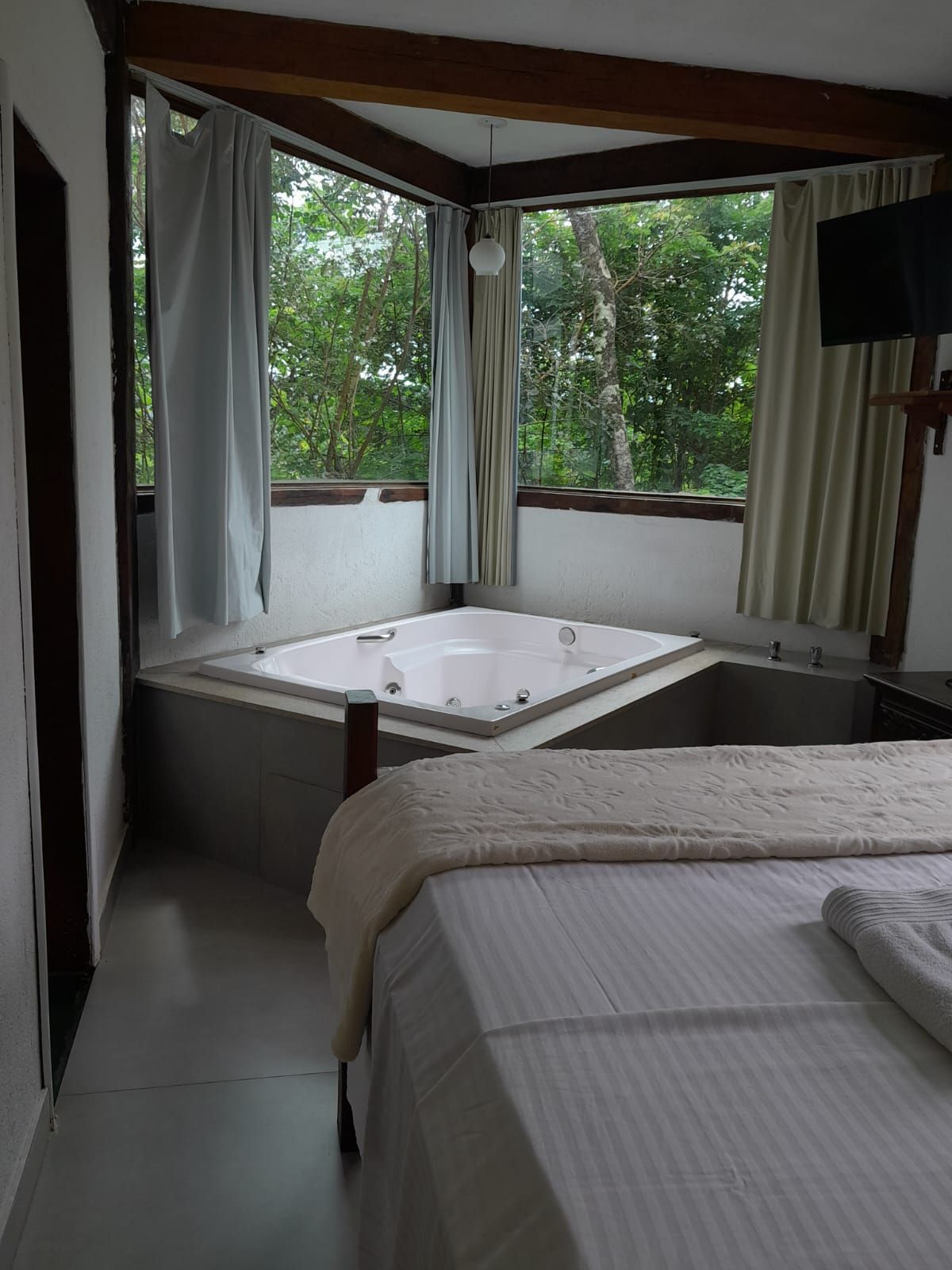 Um quarto com uma grande banheira de canto branca, janelas do chão ao teto com vista para uma floresta e uma cama em primeiro plano.