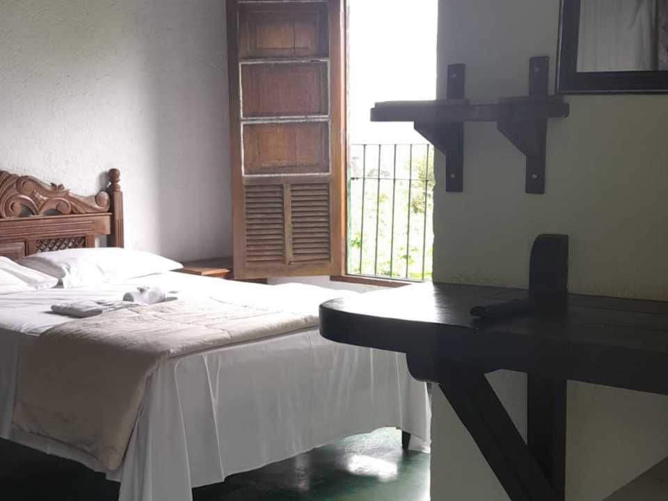 Um quarto de hotel com uma cama de madeira, lençóis brancos, uma mesa de cabeceira e uma porta de madeira aberta que dá para uma varanda.