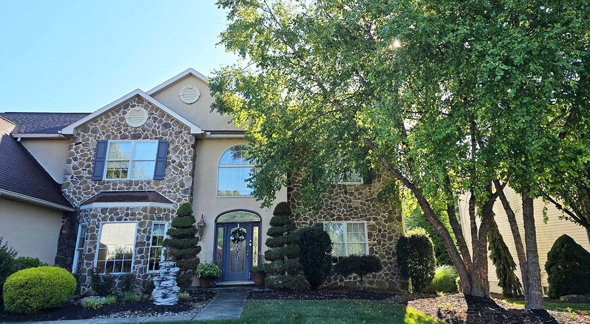 Stone-faced house with a large tree in the front yard. Blue sky above the house, sunny day.