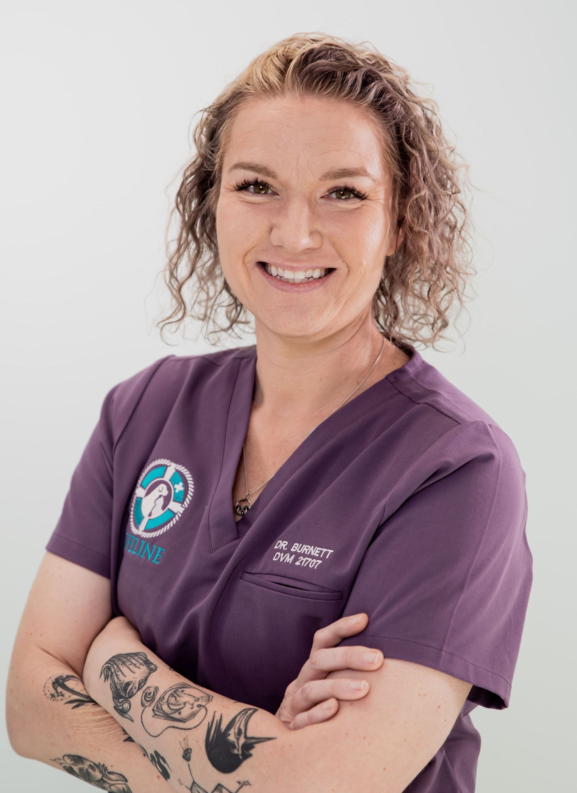 A woman in a purple scrub top is smiling with her arms crossed.