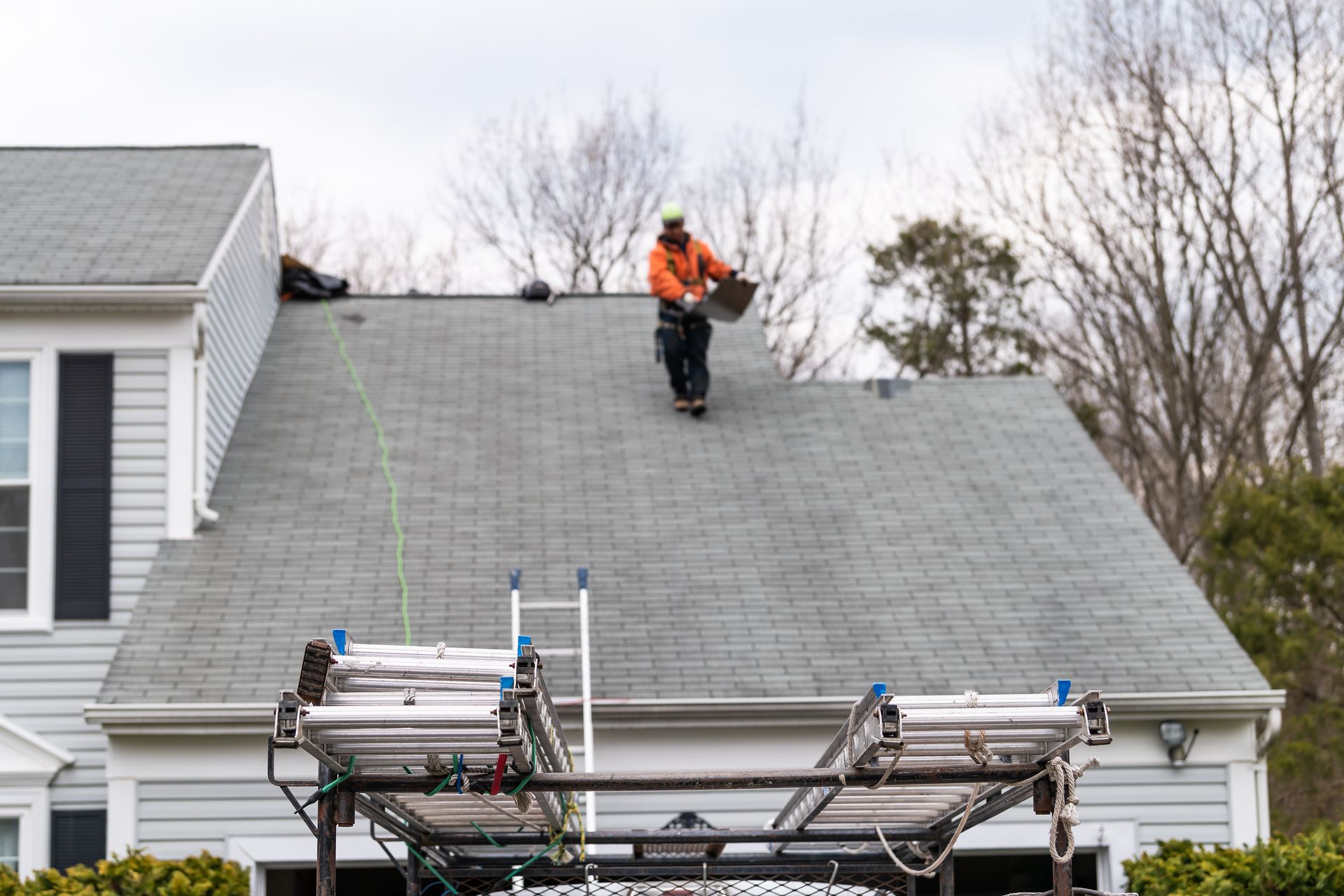Worker in orange safety gear on a gray roof above parked equipment and a house